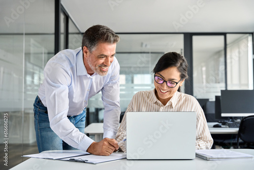 Mid aged Latin manager mentor helping Asian worker working on laptop in office. Two happy professional coworkers discussing online plan at work. Diverse business people using computer and talking.