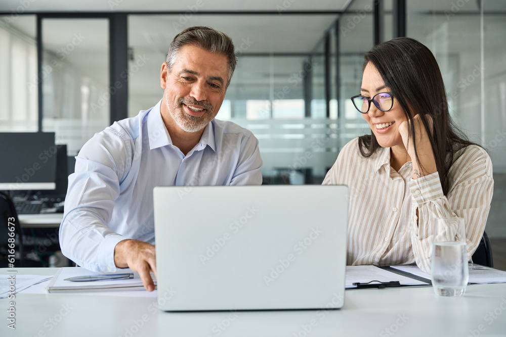 2 multicultural team coworkers, older Latin man executive and young Asian woman manager working together, happy diverse employees looking at laptop computer discussing financial corporate project. 2 multicultural team coworkers, older Latin man executive and young Asian woman manager working together, happy diverse employees looking at laptop computer discussing financial corporate project.