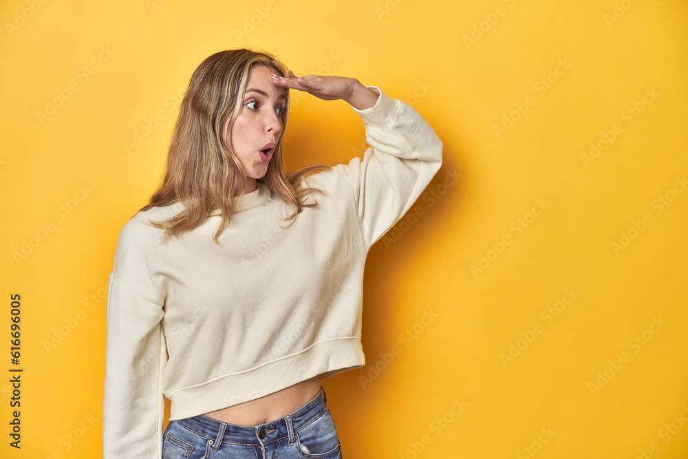 Young blonde Caucasian woman in a white sweatshirt on a yellow studio background, looking far away keeping hand on forehead. Young blonde Caucasian woman in a white sweatshirt on a yellow studio background, looking far away keeping hand on forehead.