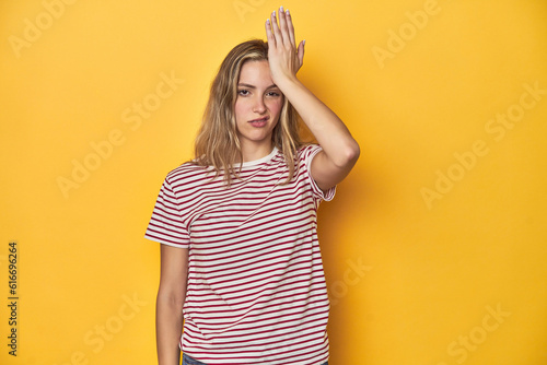 Young blonde Caucasian woman in a red striped t-shirt on a yellow background, forgetting something, slapping forehead with palm and closing eyes.