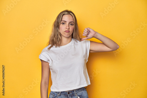 Young blonde Caucasian woman in a white t-shirt on a yellow studio background, showing thumb down, disappointment concept.