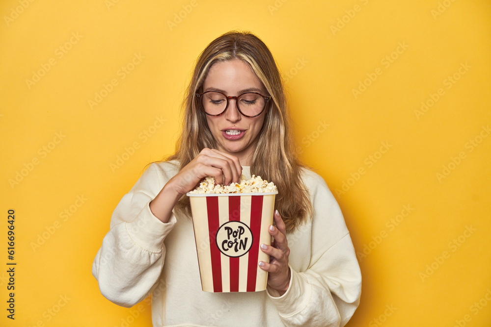 Young Caucasian woman, popcorn in hand, expressing love for cinema on a yellow backdrop.