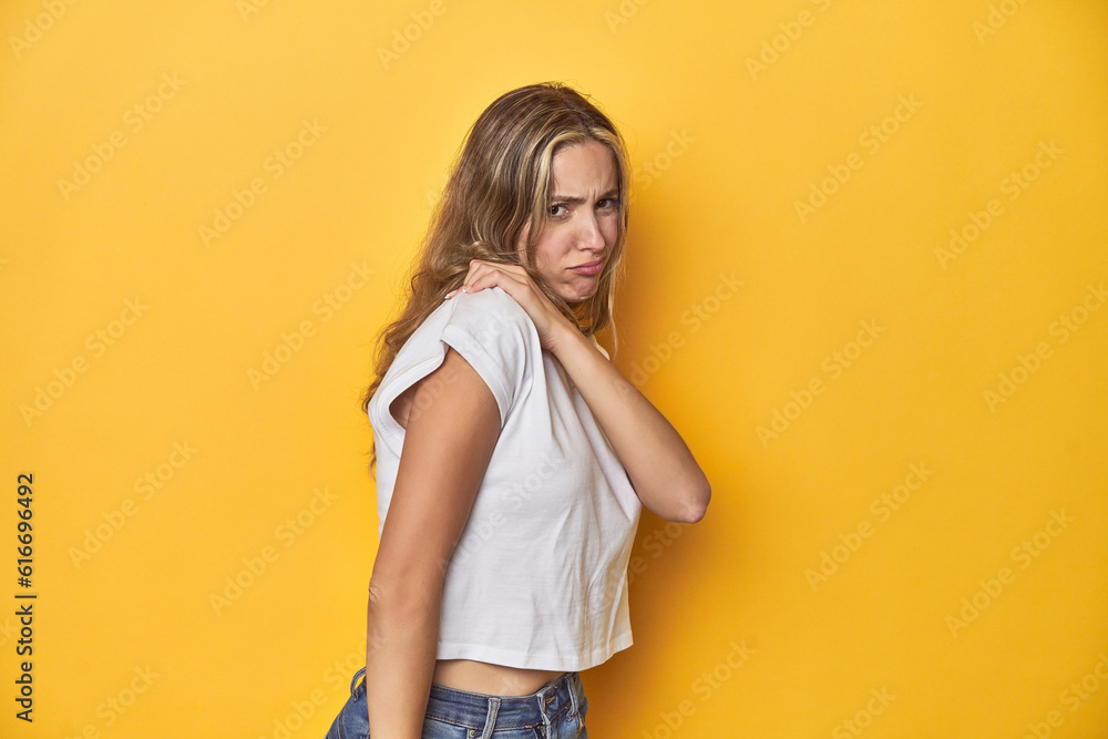 Young blonde Caucasian woman in a white t-shirt on a yellow studio background, having a shoulder pain. Young blonde Caucasian woman in a white t-shirt on a yellow studio background, having a shoulder pain.