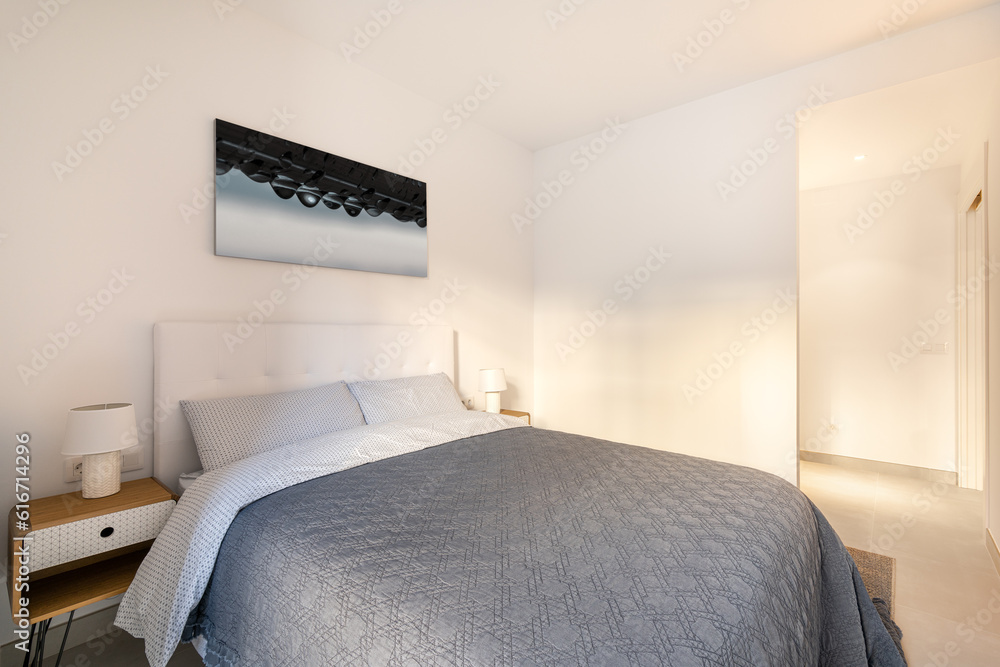 Bedroom with double bed with a picture on the wall and stylish bedside tables in light colors and an open door to the corridor. The concept of a compact hotel room. Copyspace