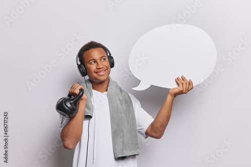 Horizontal shot of handsome dark skinned African man lifts weight hols communication bubble listens music via headphones wears t shirt and towel around neck isolated over white studio background. photo