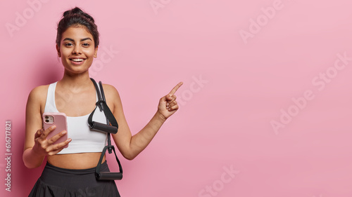 Pretty positive Indian sportswoman wears white cropped top poses with sport equipment holds smartphone points index finger aside on blank space advertises something isolated over pink background