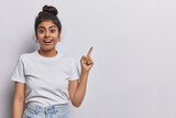 Horizontal shot of pretty surprised cheerful young woman pointing to empty copy space advertises product or tells about awesome offer dressed in casual clothing isolated over white background