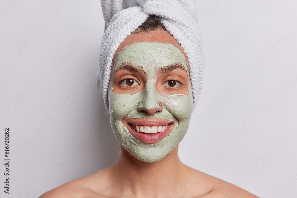 Joyous female model exudes happiness applies nourishing facial mask for skin treatment smiles broadly poses with bath towel wrapped on head isolated over white background. Pampering procedures concept Joyous female model exudes happiness applies nourishing facial mask for skin treatment smiles broadly poses with bath towel wrapped on head isolated over white background. Pampering procedures concept
