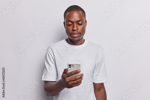 Waist up shot of serious dark skinned man concentrated in smartphone dressed in casual t shirt gets message or reads news in internet isolated over white background. People and technology concept