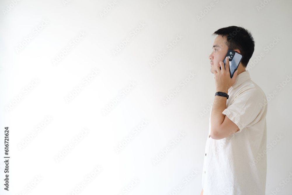 A young Asian man wearing a beige shirt and a smartwatch on his left wrist is on the phone while looking to his right side or left side of the camera. Isolated white background. A young Asian man wearing a beige shirt and a smartwatch on his left wrist is on the phone while looking to his right side or left side of the camera. Isolated white background.