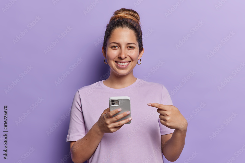 Horizontal shot of pretty young female model holding cellphone advertising new trendy application points at modern gadget smiles toothily wears casual t shirt isolated over purple background Horizontal shot of pretty young female model holding cellphone advertising new trendy application points at modern gadget smiles toothily wears casual t shirt isolated over purple background