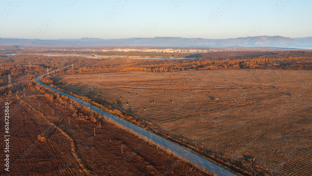Autumn aerial photograph of the road among the fields. Tractors mow hay in the fields. A small town is in the distance. Beautiful rural landscape. Travel through the countryside.