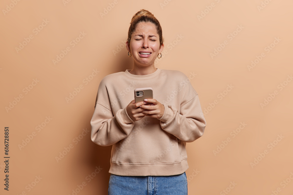 Depressed young woman standing with cell phone in hands and crying having bad news cries from despair wears casual jumper jeans isolated over brown background. Negative human emotions and technology Depressed young woman standing with cell phone in hands and crying having bad news cries from despair wears casual jumper jeans isolated over brown background. Negative human emotions and technology