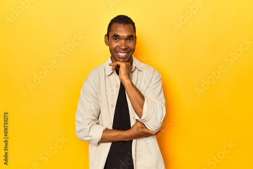 African American man with shirt, yellow studio, smiling happy and confident, touching chin with hand.