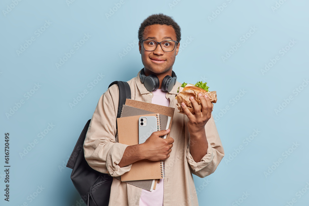 Education and lifestyle concept. Handsome dark skinned male student eats sandwich after day of studying holds spiral notepads carries rucksack dressed casually isolated over blue background. Education and lifestyle concept. Handsome dark skinned male student eats sandwich after day of studying holds spiral notepads carries rucksack dressed casually isolated over blue background.