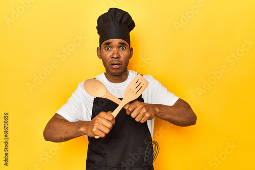 African American cook holding wooden spoon and fork in a yellow studio doing a denny gesture photo