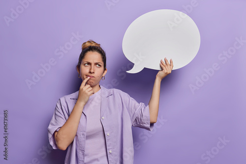 Canvas Print Displeased European woman has hair combed in bun with thoughtful expression holds communication bubble for your advertisement purses lips dressed casually isolated over purple background