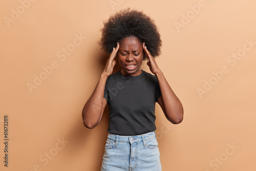 Black curly haired woman rubs her temples squinting in pain as debilitating headache or migraine takes hold overwhelmed by exhaustion wears black t shirt and jeans isolated over brown background
