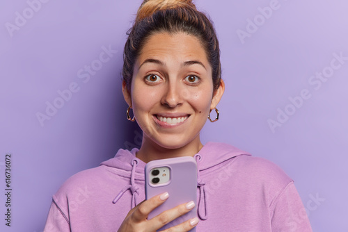 Image of smiling woman chatting in app holding smartphone looking at camera happilly texting message smiles toothily dressed in casual sweatshirt isolated over purple background. Online communication photo