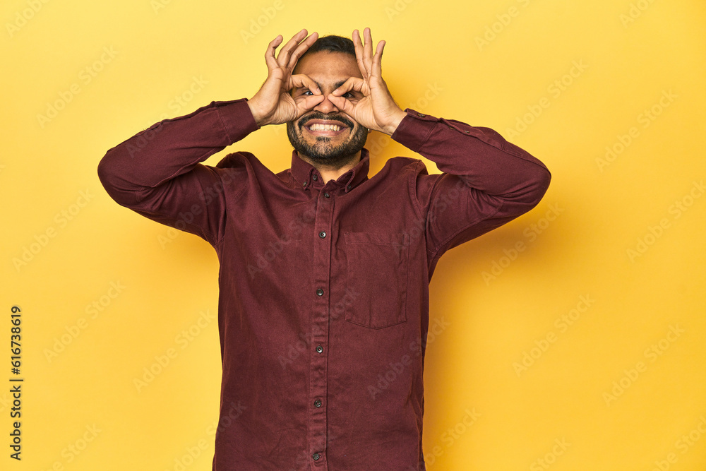 Casual young Latino man against a vibrant yellow studio background, showing okay sign over eyes Casual young Latino man against a vibrant yellow studio background, showing okay sign over eyes