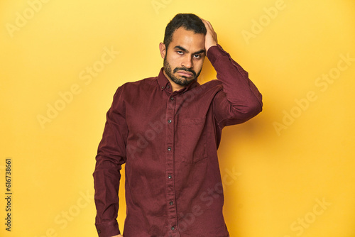 Casual young Latino man against a vibrant yellow studio background, tired and very sleepy keeping hand on head.