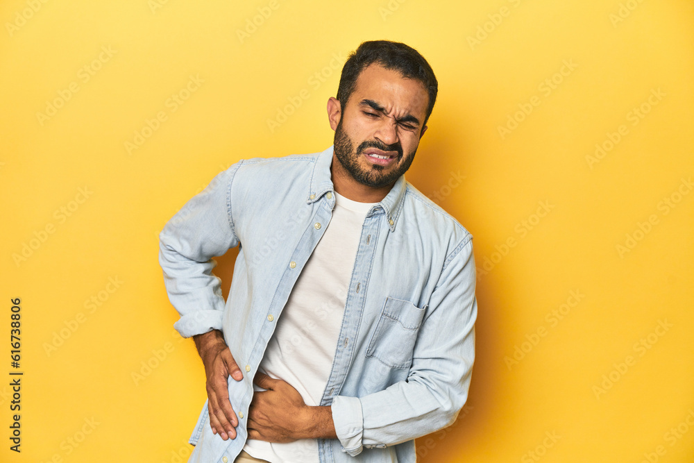 Casual young Latino man against a vibrant yellow studio background, having a liver pain, stomach ache. Casual young Latino man against a vibrant yellow studio background, having a liver pain, stomach ache.