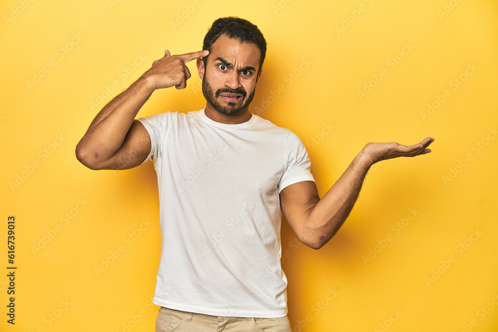 Casual young Latino man against a vibrant yellow studio background, holding and showing a product on hand. Casual young Latino man against a vibrant yellow studio background, holding and showing a product on hand.