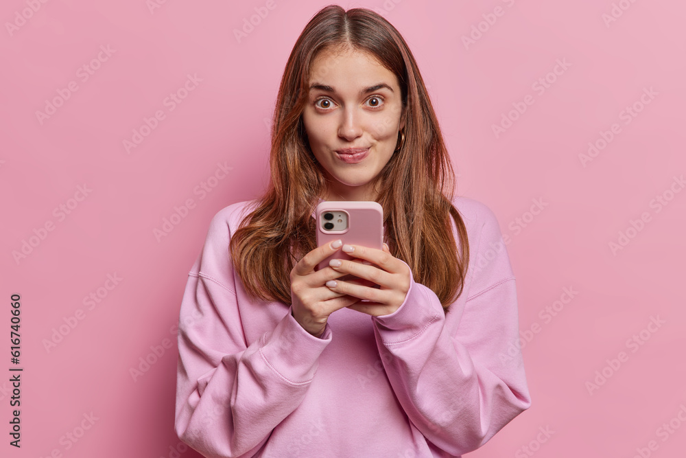Surprised young European woman with intriguing expression holds smartphone chats online browses internet dressed in casual pullover isolated over pink background. People reactions technology concept Surprised young European woman with intriguing expression holds smartphone chats online browses internet dressed in casual pullover isolated over pink background. People reactions technology concept