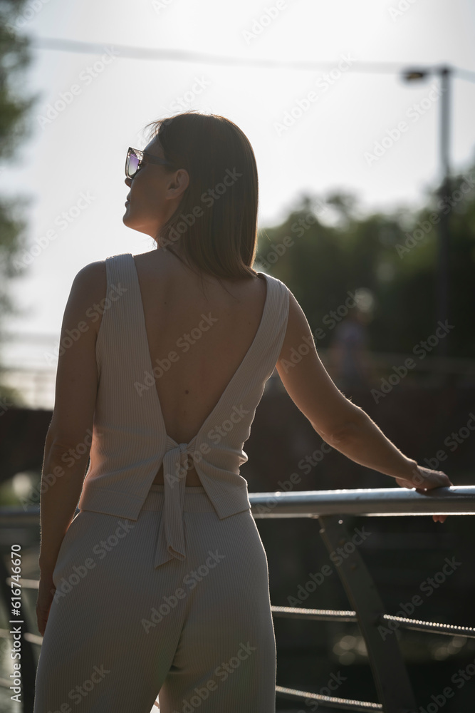 Summer urban portrait of a young brunette woman in sunglasses. The portrait was taken during the golden hour under backlight.