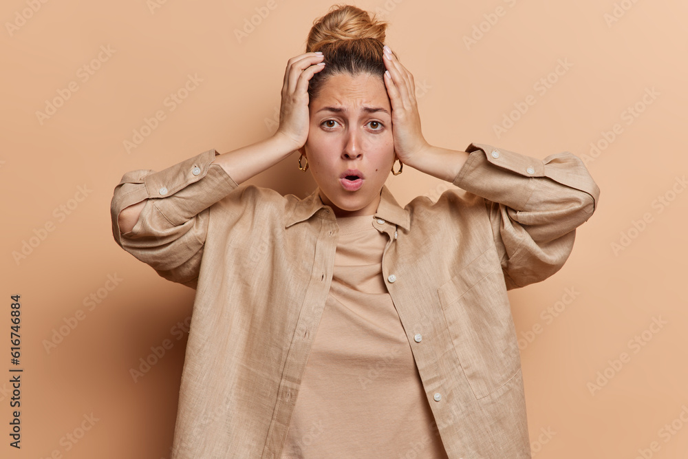 Shocked scared woman keeps hands on head says omg has fair hair combed in bun wears shirt isolated over brown background. Speechless female model reacts to something very shocking and horrible Shocked scared woman keeps hands on head says omg has fair hair combed in bun wears shirt isolated over brown background. Speechless female model reacts to something very shocking and horrible