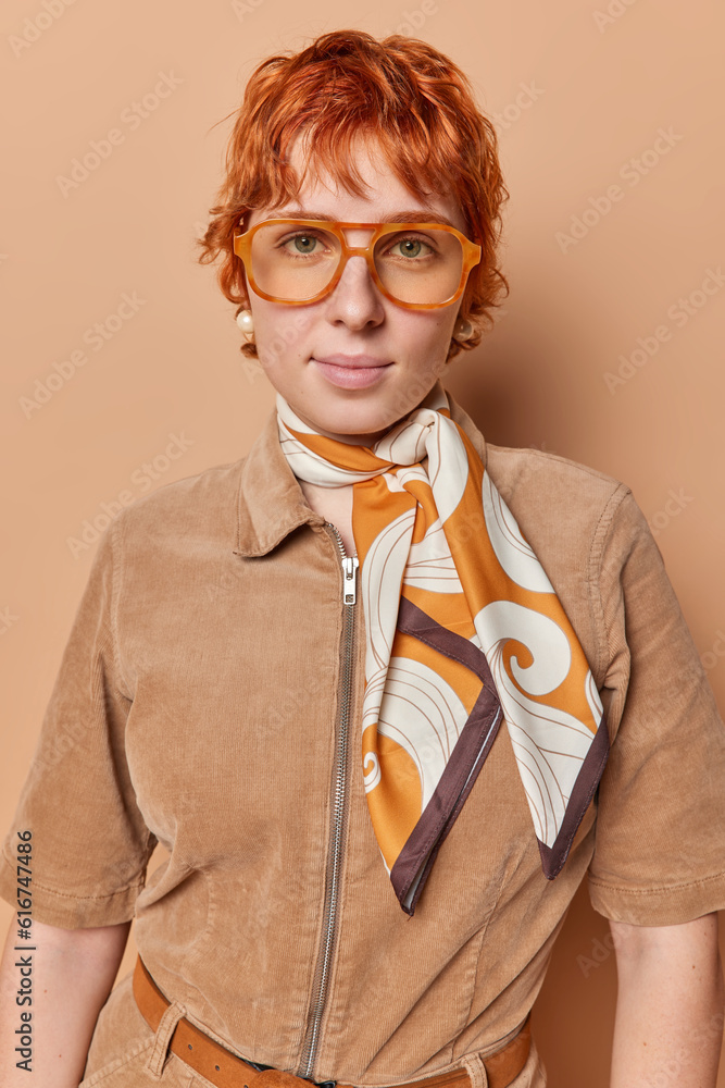 Φωτογραφία Mesmerizing redhead woman looks attentively at camera dressed in fashionable clothes wears transparent eyeglasses has serious expression isolated over brown background