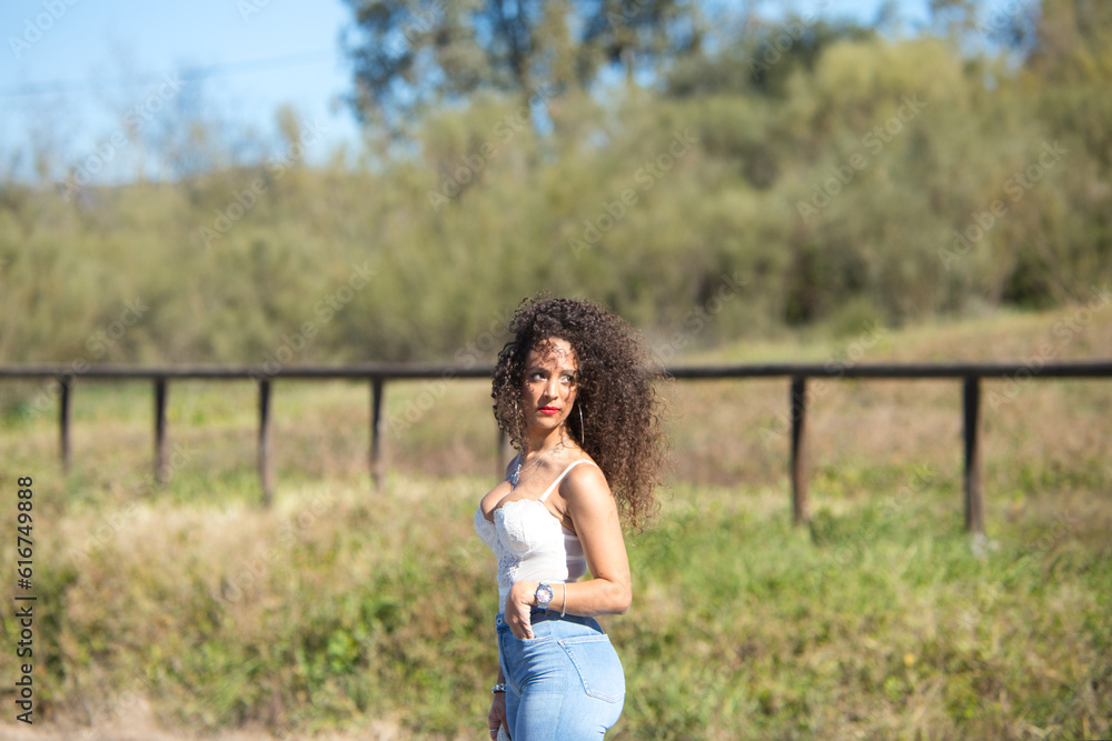 Spanish woman with dark curly hair wearing jeans and white top and red lips is walking in the countryside on a sunny day. Spanish woman with dark curly hair wearing jeans and white top and red lips is walking in the countryside on a sunny day.