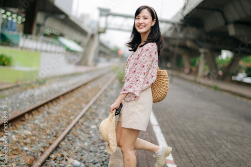 young asian woman traveler with weaving basket happy smiling looking to a camera beside train railway. Journey trip lifestyle, world travel explorer or Asia summer tourism concept. © Jirawatfoto