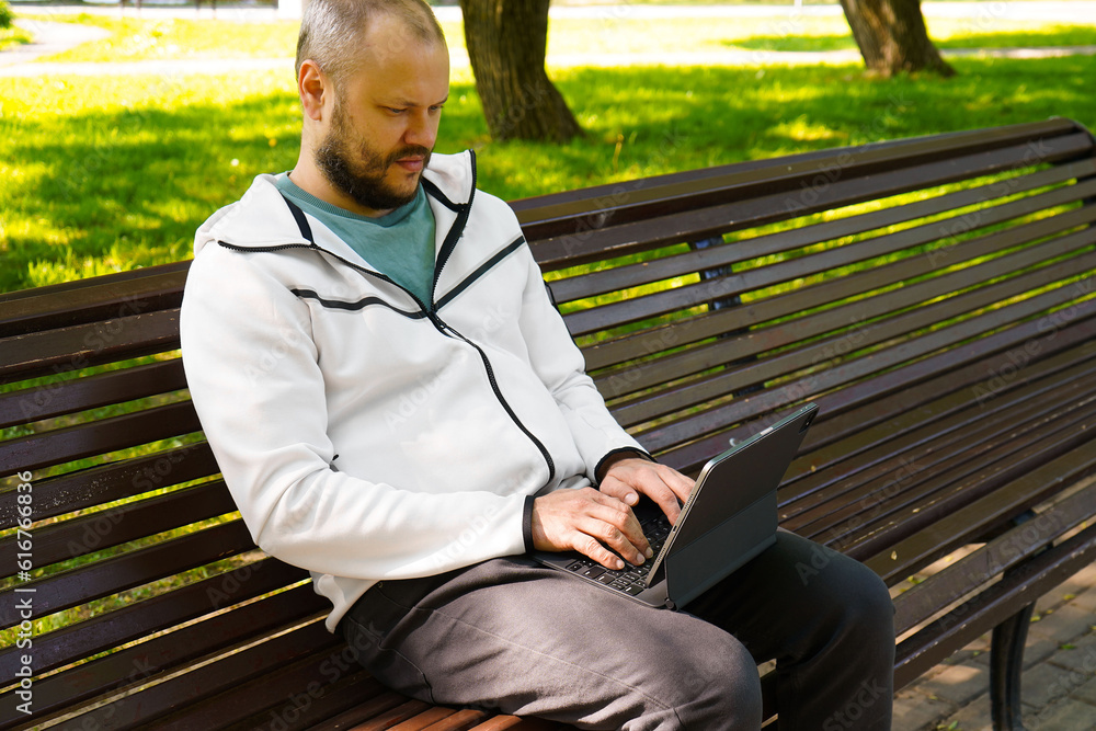 Fototapeta premium Man freelancer holding black tablet pc with mock up green desktop screen sitting on a bench in the park