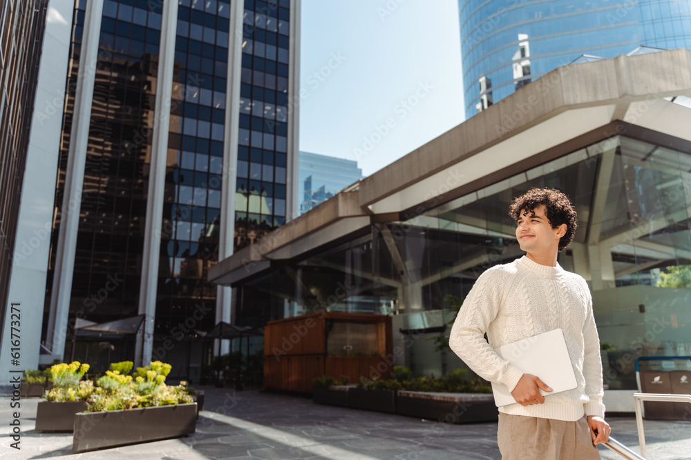 Portrait of confident young businessman holding laptop, standing on urban street looking away Portrait of confident young businessman holding laptop, standing on urban street looking away