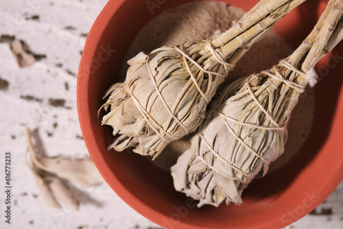 Overhead view of two smudge sticks with White Sage (Salvia apiana) and medicinal pouch in a clay bowl on old white wood