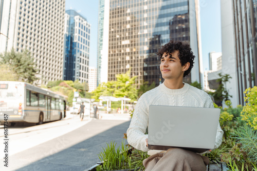 Portrait of attractive smiling latino man using laptop, working on urban street, technology concept
