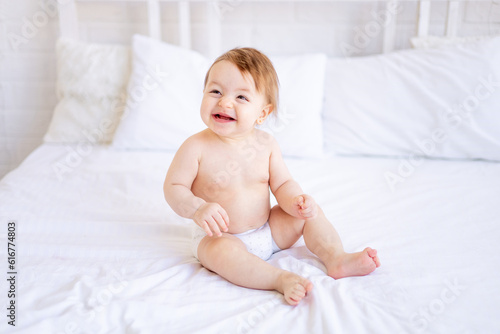 a joyful baby girl of six months on a bed in diapers smiles, a small child on a cotton bed at home, the concept of care and hygiene