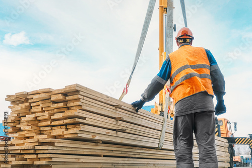 A slinger unloads wooden planks outdoors on a summer day. A worker in a hard hat and high-visibility vest stacks lumber. Industrial background with copy space.
