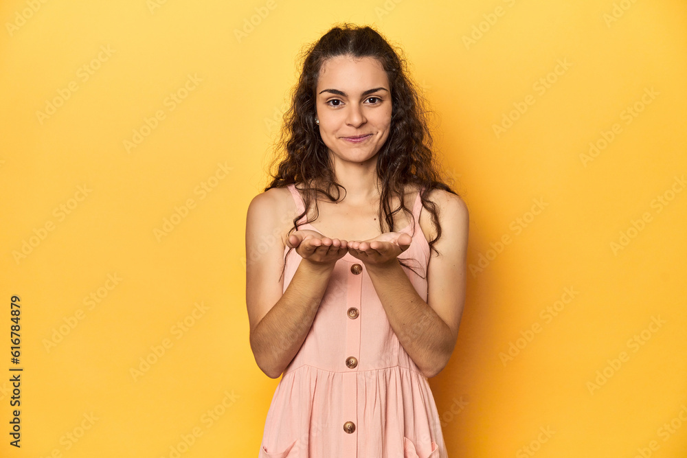 Young Caucasian woman, yellow studio background, holding something with palms, offering to camera. Young Caucasian woman, yellow studio background, holding something with palms, offering to camera.