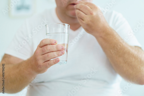 Unwell caucasian man got a sickness, oversize fat man taking a pills and drinking water in a drinking glass after wake up in morning.