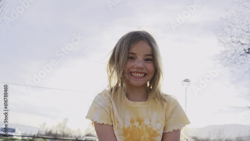 Smiling little Caucasian girl looks at camera love expression outdoors on sunny day. Cute giggling blonde kid poses naughty for video sitting in park. Concept of childhood in family and leisure time. photo