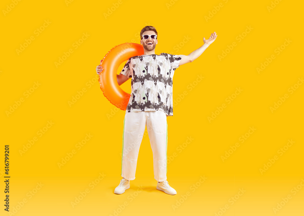 Smiling happy guy in sunglasses holding inflatable rubber ring. Full length portrait of excited young man wearing summer casual clothes standing on isolated yellow studio background Smiling happy guy in sunglasses holding inflatable rubber ring. Full length portrait of excited young man wearing summer casual clothes standing on isolated yellow studio background