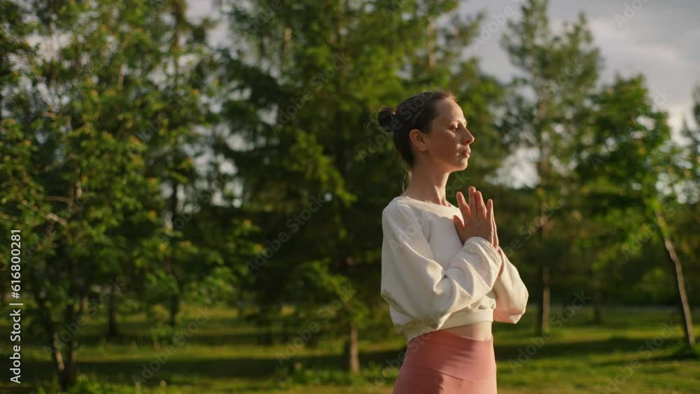 Side view of relaxed yoga woman clasping hands on chest making Namaste position standing on background of trees. Tranquil young woman meditating outdoors at sunrise. Calm lady feeling calmness harmony Side view of relaxed yoga woman clasping hands on chest making Namaste position standing on background of trees. Tranquil young woman meditating outdoors at sunrise. Calm lady feeling calmness harmony