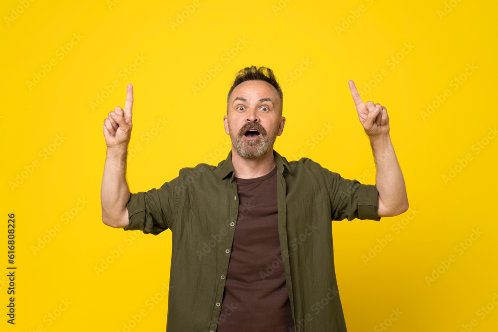 50s age excited handsome mature bristle man in green shirt, pointing upwards with finger, having great idea, isolated on yellow background. Mustach man pointing above by finger shocked with open mouth 50s age excited handsome mature bristle man in green shirt, pointing upwards with finger, having great idea, isolated on yellow background. Mustach man pointing above by finger shocked with open mouth