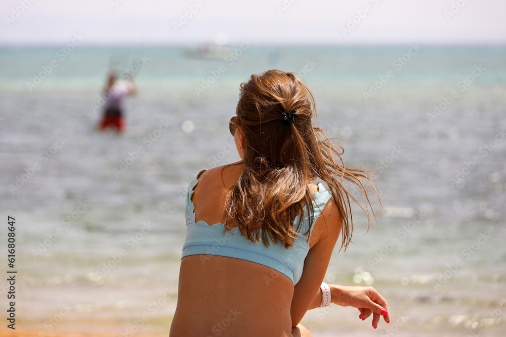 Naklejka premium Girl in swimsuit sitting on sea beach and looking at a man engaged in parasailing. Relaxation on summer resort