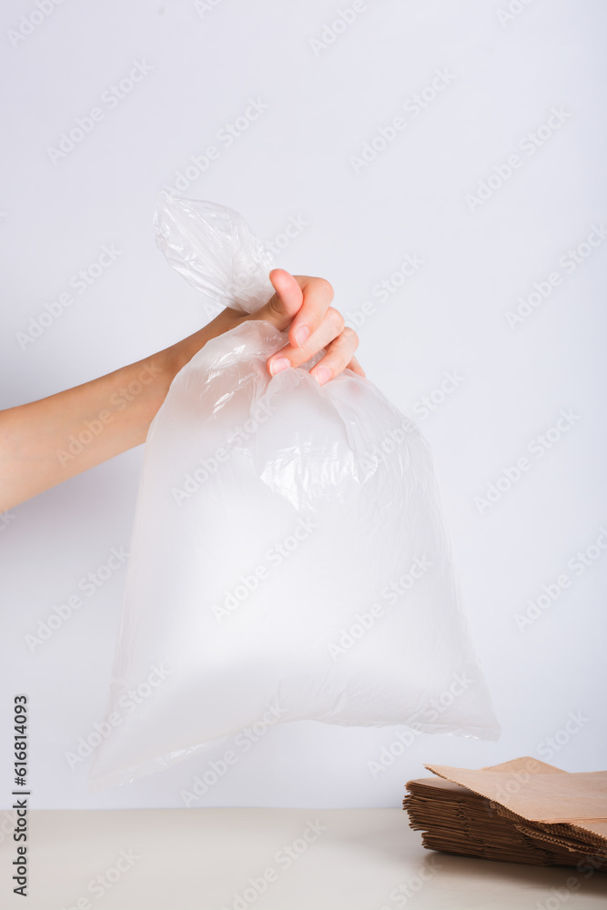 Naklejka premium Hand holding an empty transparent plastic bag and a stack of eco-friendly paper bags on the table
