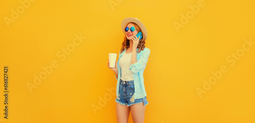 Happy smiling young woman calling on smartphone wearing straw hat, shorts on yellow background