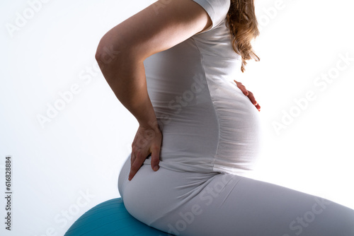 Pregnant woman with baby bump on gymnastic ball feels back pain and holds hand on her aching back. Final month of pregnancy - week 36. Side view. White background. Bright shot.