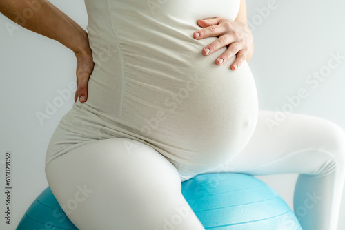 Mother with heavily pregnant baby bump sitting on gym ball treats back pain and holds hand on her aching back. Side view. White background. Bright shot.